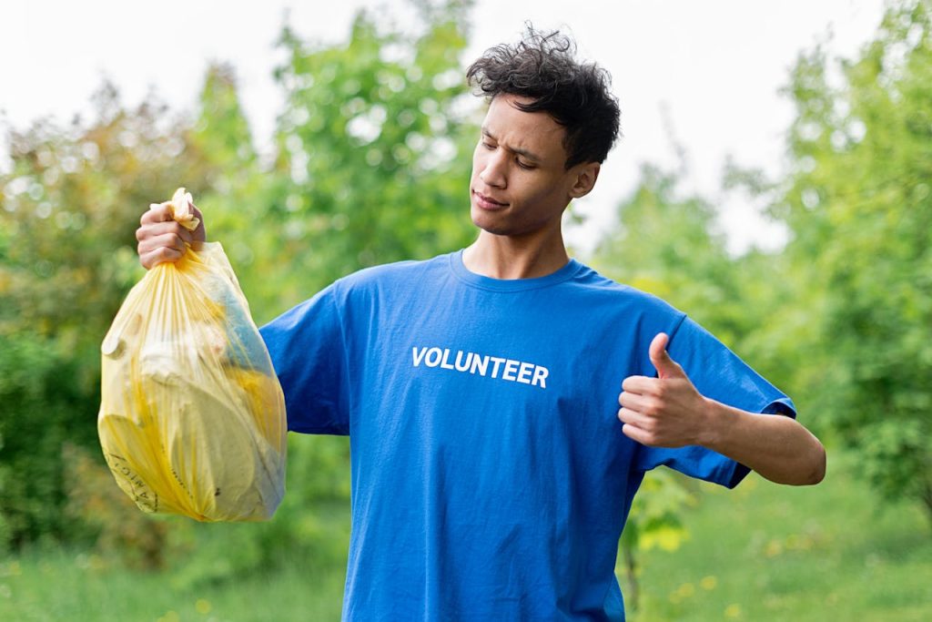 a-man-holding-a-bag-of-plastics-8543570 Young volunteer in a blue shirt holding a bag of recyclables and giving a thumbs up outdoors.